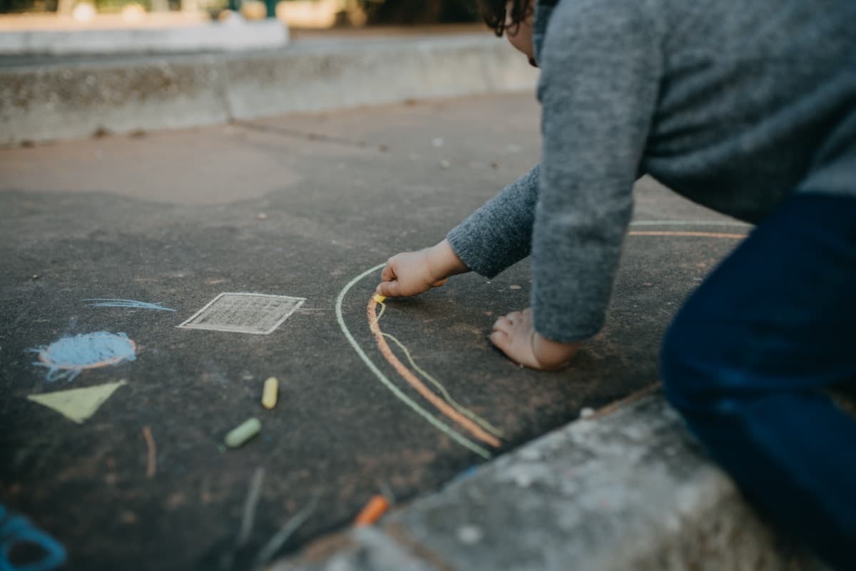 Ein Kind malt mit Kreide einen bunten Regenbogen auf den Asphalt als Sinnbild für ambulante Hilfe, die im Alltag von Kindern ansetzt