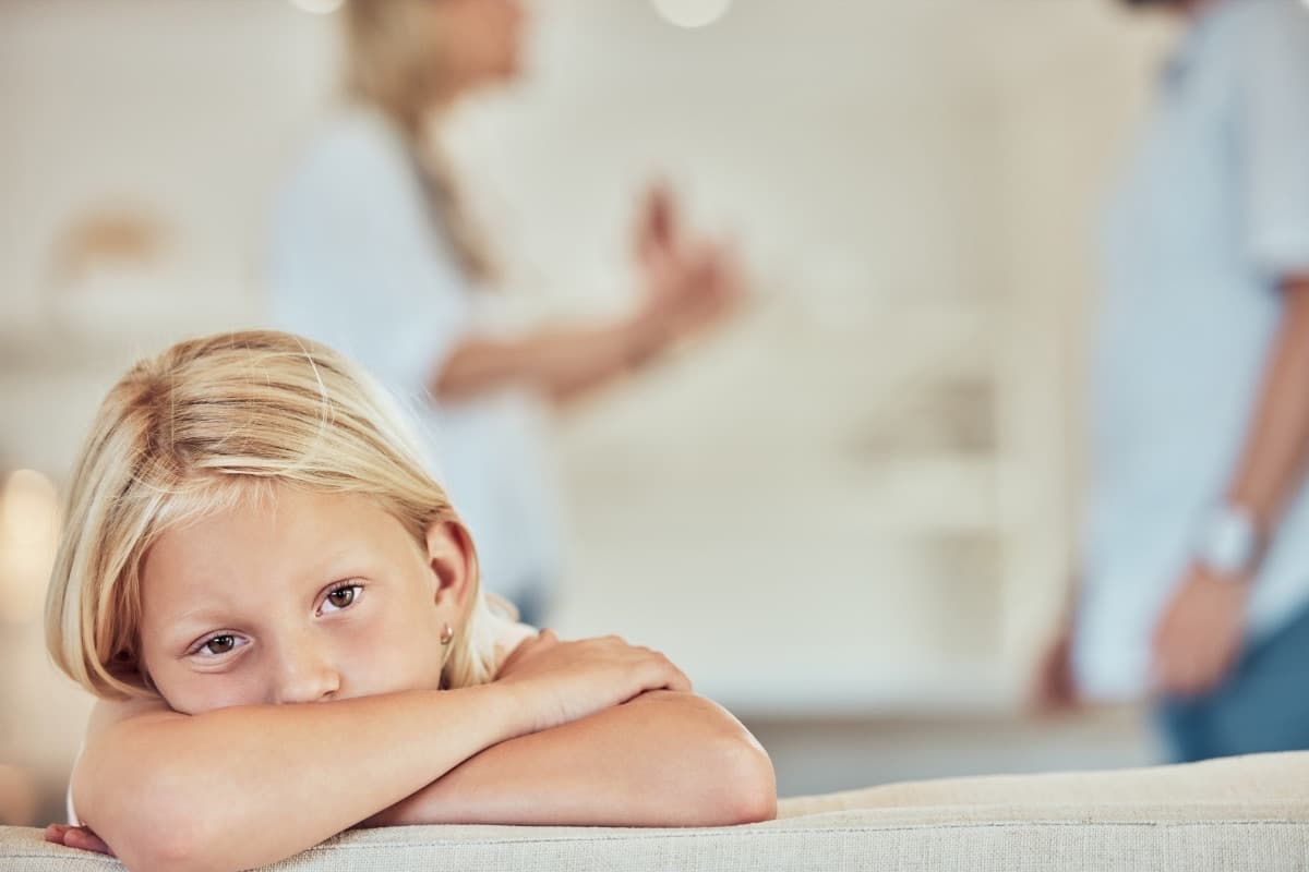 Mädchen lehnt nachdenklich auf einer Sofalehne, im Hintergrund unscharf die Eltern