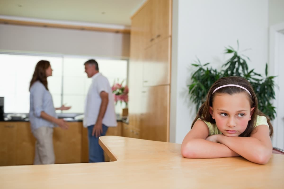 Nachdenkliches Mädchen am Tisch, im Hintergrund unscharf die Eltern in der Küche