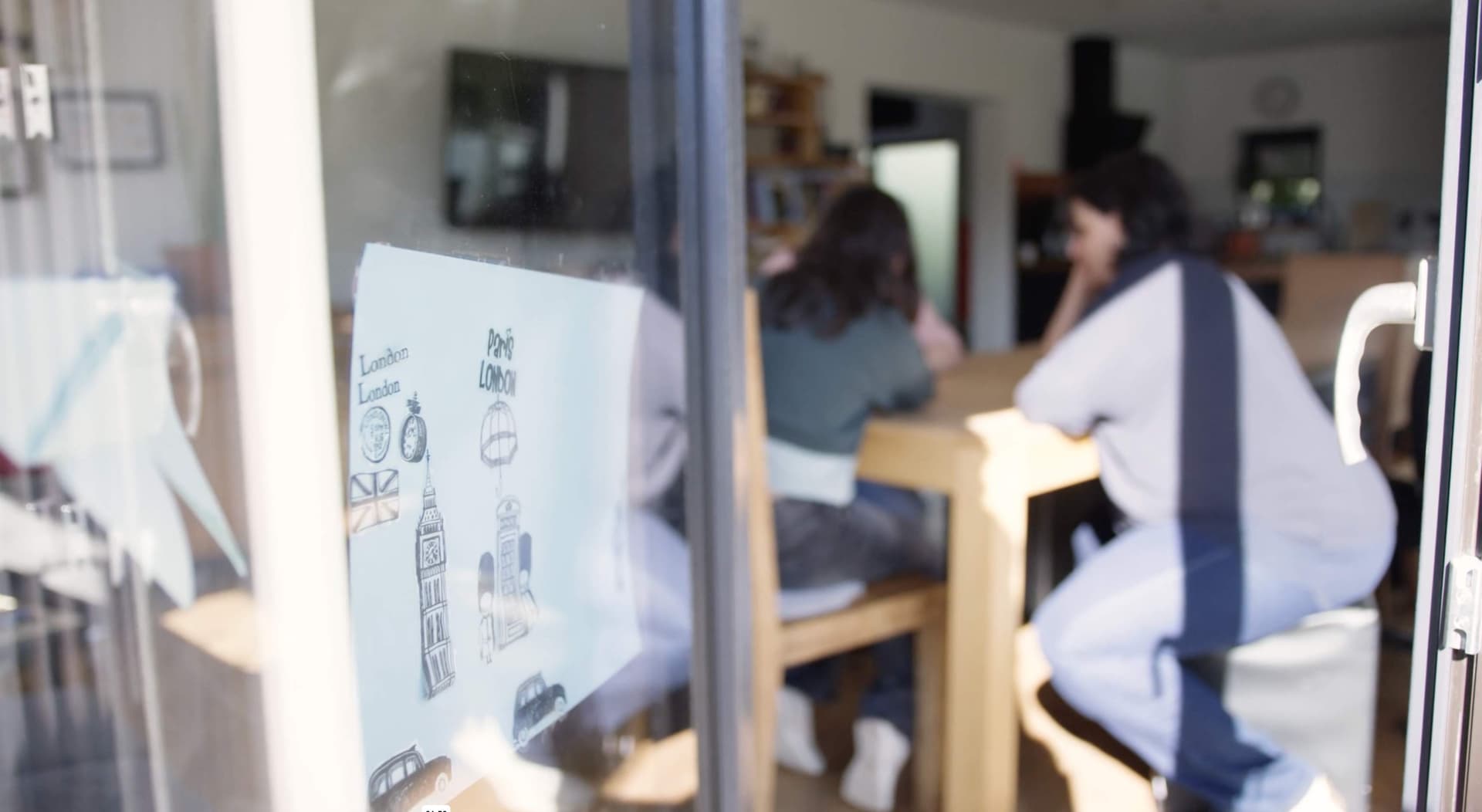 Zwei Jugendliche sitzen am Esstisch einer Wohngruppe, gesehen durch eine Glastür mit Kinderzeichnung, als Symbol für Alltag, Nähe und ein echtes Zuhause